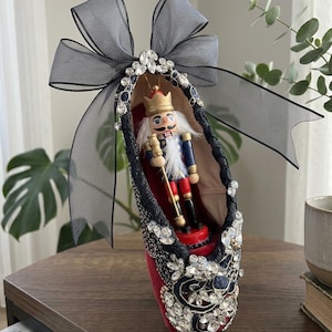 May include: A red ballet slipper ornament decorated with black lace, clear rhinestones, and a large sheer black ribbon bow. Inside the shoe sits a small wooden nutcracker king figure wearing a gold crown, blue jacket, and red trousers.