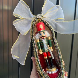 May include: A decorative shoe ornament featuring a miniature nutcracker soldier in a green and red uniform. The shoe is lined with red fabric, trimmed with gold tinsel and plaid ribbon, and topped with a large white sheer bow with gold edges.