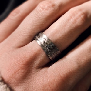 Könnte beinhalten: Ein silberner Ring mit einem strukturierten Design. Der Ring wird an einem Finger getragen.