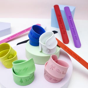Può includere: Una collezione di braccialetti e portabicchieri in silicone colorati. I braccialetti sono rossi, viola e rosa, con la scritta "Be happy every day". I portabicchieri sono blu, bianchi, verdi, gialli e rosa, con vari disegni e testo.