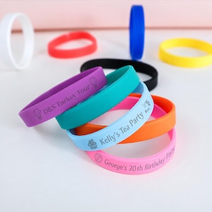 Könnte beinhalten: Eine Sammlung bunter Silikonarmbänder in Weiß, Rot, Blau, Gelb, Schwarz, Lila, Türkis, Hellblau, Orange und Pink. Einige Bänder sind mit Text und Symbolen bedruckt, darunter D&S Turkey Tour, Kelly's Tea Party und George's 20th Birthday Party.