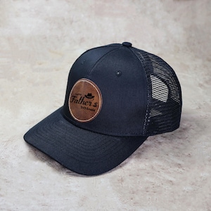 Może przedstawiać: Czarna czapka typu trucker z zakrzywionym daszkiem i siatkowym tyłem. Brązowa skórzana naszywka z przodu z napisem "Happy Father's Day Gerald" w okrągłym wzorze, z grafiką kapelusza.