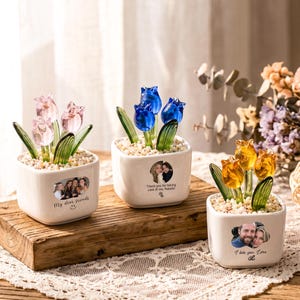 Op de afbeelding: Drie vierkante witte keramische plantenbakken met glazen tulpen in roze, blauw en amber. Elke pot is aan de voorzijde voorzien van een aangepaste foto en tekst, gevuld met kleine lichtgekleurde steentjes en groene glazen bladeren.
