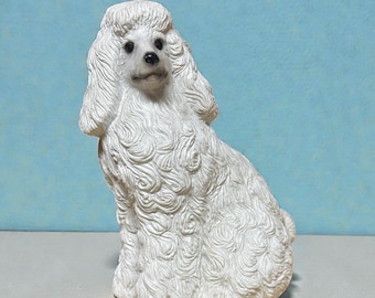 Könnte beinhalten: Eine weiße Pudelfigur in sitzender Position. Der Hund hat ein lockiges Fell und ein detailliertes Gesicht mit schwarzen Augen und einer schwarzen Nase. Die Figur steht auf einer weißen Oberfläche mit hellblauem Hintergrund.