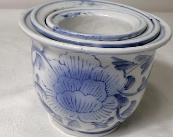 May include: A white ceramic planter with a blue floral design. The planter has a round shape with a wide rim and a matching set of three nested saucers. The floral pattern features a large flower and leaves.