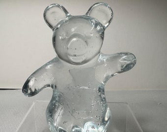 Könnte beinhalten: Eine klare Glasbärenfigur mit ausgestreckten Armen und sichtbaren Luftblasen. Der Bär hat abgerundete Ohren und einen glatten, transparenten Körper. Die Figur wird auf einem transparenten, rechteckigen Ständer ausgestellt.