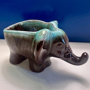 May include: A ceramic planter shaped like an elephant with a glossy finish. The body features a deep brown base color that transitions into a vibrant turquoise glaze along the top rim and ears, with a trunk extending forward from the front.