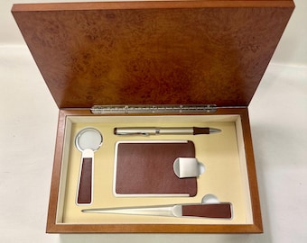 May include: A wooden desk set with a hinged lid, containing a pen, letter opener, magnifying glass, and a business card holder. The set has a brown and beige color scheme, with a wood grain finish on the box and brown leather accents.