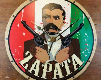 May include: Round wooden wall clock featuring a portrait of Emiliano Zapata holding two revolvers. The clock face has a red, white, and green background, with the word "ZAPATA" in bold letters. The clock hands are gold.
