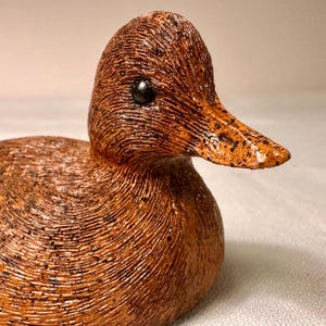 May include: A detailed, handcrafted duck figurine. The duck features a textured, brown body with black speckles and a contrasting orange beak. The eye is a small, black bead. The figurine is a decorative item.