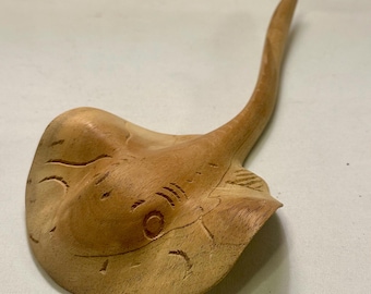 May include: A wooden sculpture of a stingray, featuring a light brown color with carved details. The sculpture has a curved tail and detailed facial features, including eyes and gill slits. The wood grain is visible.