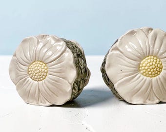 May include: Two ceramic drawer knobs shaped like white flowers with pale yellow centers. The petals have a glossy finish, while the sides of the knobs feature a textured, dark green leaf pattern that wraps around the base of each floral shape.
