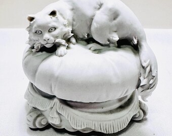Könnte beinhalten: Eine weiße Keramik-Katzenfigur, die auf einem dekorativen Kissen liegt. Die Katze hat detailliertes Fell und Gesichtszüge, mit dem Schwanz um das Kissen gerollt. Das Kissen hat einen gerüschten Rand und steht auf einem dekorativen Sockel.