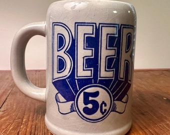 May include: A cream-colored ceramic beer mug with a prominent crackle glaze finish. The front features bold blue text that reads BEER above a circular graphic containing the number 5 and a cent symbol, all set against a white background.