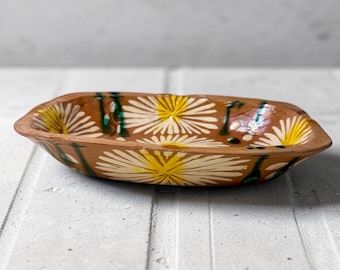 May include: A shallow, rectangular terracotta bowl with rounded corners, featuring a hand-painted pattern of white and yellow daisy-like flowers and thin green stems against a warm, earthy brown clay background with a glossy finish.