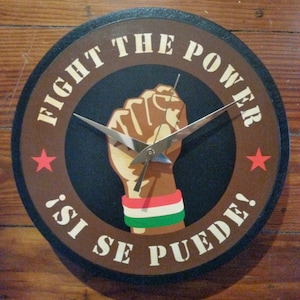 May include: Round wall clock with a brown border and a black center. The clock face features a raised fist with a red, white, and green wristband. The words "FIGHT THE POWER" and "¡SI SE PUEDE!" are printed on the clock face.