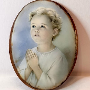 May include: Oval portrait of a child with blonde curly hair, blue eyes, and clasped hands. The child is wearing a white garment against a light blue background. The portrait is framed in a dark brown, aged wooden frame.