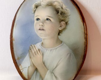 May include: Oval portrait of a child with blonde curly hair, blue eyes, and clasped hands. The child is wearing a white garment against a light blue background. The portrait is framed in a dark brown, aged wooden frame.