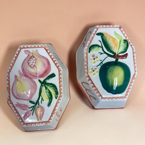 May include: Two hexagonal ceramic wall plaques featuring hand-painted fruit designs. One plaque displays pink pomegranates with green leaves, while the other shows a green apple with yellow leaves and small white flowers, both with beaded borders.