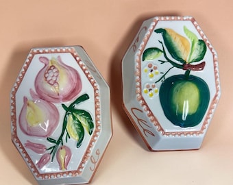 May include: Two hexagonal ceramic wall plaques featuring hand-painted fruit designs. One plaque displays pink pomegranates with green leaves, while the other shows a green apple with yellow leaves and small white flowers, both with beaded borders.