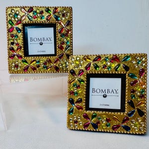May include: Two square picture frames with a gold glitter finish, decorated with multicolored faceted jewels in floral patterns. Each frame features a white insert with the text BOMBAY and 2 x 2 inches printed in black, centered within the frame.