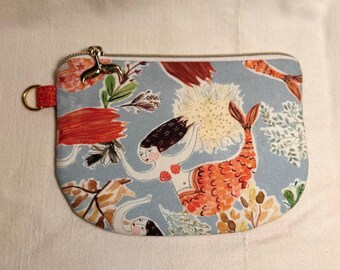 May include: A small, rectangular coin purse with a light blue background featuring a mermaid pattern. The mermaids have orange tails and dark hair. The purse has a red loop and a gold zipper pull in the shape of a deer.
