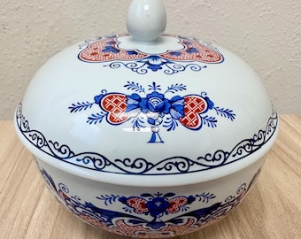 Könnte beinhalten: Eine weiße Keramikschale mit Deckel, abgerundeter Form und einem knopfförmigen Griff. Die Oberfläche ist mit detaillierten blauen und orangefarbenen Blumenmustern, symmetrischen Schnörkeln und geometrischen Motiven in horizontalen Bändern verziert.