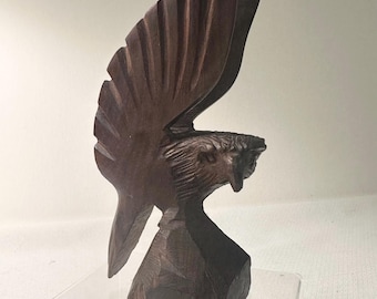 Könnte beinhalten: Handgeschnitzte Holzskulptur eines fliegenden Adlers. Der Adler hat ausgebreitete Flügel und einen detaillierten Kopf, der die Textur des Holzes zeigt. Die Skulptur ist dunkelbraun und steht auf einem transparenten, rechteckigen Sockel.