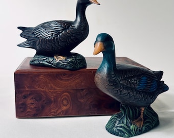 May include: Two duck figurines with textured feathers and orange beaks. One duck is dark brown and positioned on a wooden box, while the other is dark blue with green accents and stands on a base of sculpted grass, both featuring detailed plumage.
