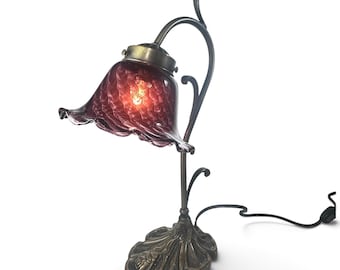 Könnte beinhalten: Eine Tischleuchte mit einem tiefvioletten, gerüschten Glasschirm mit strukturierter Oberfläche. Die Lampe hat einen geschwungenen, dunkel bronzefarbenen Metallschaft und einen dekorativen Sockel mit Blattmuster sowie ein schwarzes Stromkabel.