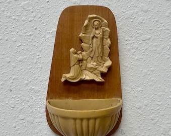 Könnte beinhalten: Ein Weihwasserbecken zur Wandmontage mit einem hellbeigen Relief aus geformtem Material, das eine kniende und eine stehende Figur in Gewändern vor einer Felsformation zeigt, montiert auf einer dunkelbraunen Holzplatte mit halbrunder Schale.