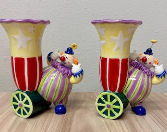 May include: Two ceramic clown-themed vases featuring a yellow top with white stars, red and white vertical stripes, and a base with green wheels. Each clown wears a purple and green striped outfit with a ruffled collar and a small yellow flower.