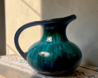 May include: A ceramic pitcher featuring a rounded body and a curved handle. The surface is finished with a glossy glaze that transitions from a deep, dark navy blue at the base to a vibrant, bright teal green toward the top, creating a smooth gradient.