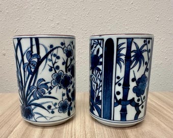 Könnte beinhalten: Zwei zylindrische Keramikbecher mit weißem Untergrund und detaillierten, handgemalten blauen botanischen Illustrationen. Der linke Becher zeigt florale Muster, während der rechte Becher segmentierte Bambusstängel und Blattwerk aufweist.