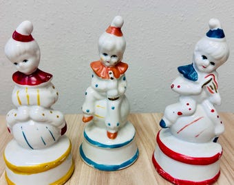 Könnte beinhalten: Drei Porzellan-Clownfiguren, die jeweils auf einem Ball auf einem runden Sockel sitzen. Der linke Clown trägt Rot und Gelb, der mittlere Clown Orange und Blau, und der rechte Clown Blau und Rot. Jede Figur hat ein bemaltes Gesicht.