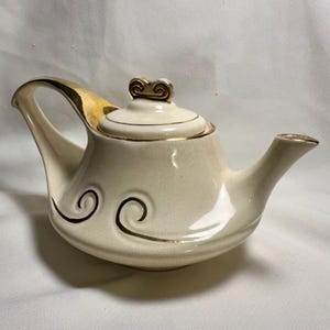 May include: Cream-colored ceramic teapot with gold accents. The teapot features a curved handle, a spout, and a lid with a decorative finial. Black swirl designs adorn the body, and gold trim outlines the edges.