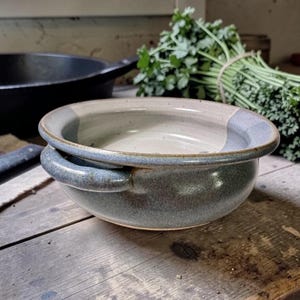 May include: A round ceramic bowl with two small side handles. The exterior features a mottled blue-grey glaze, while the interior is finished in a smooth, light cream-colored glaze. The bowl has a wide, rounded rim and a deep, circular basin.