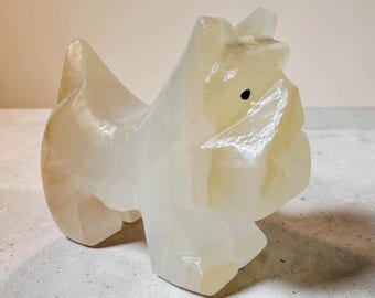 Könnte beinhalten: Eine polierte, durchscheinende cremeweiße Steinstatuette in Form eines kleinen Hundes. Die Skulptur zeichnet sich durch glatte, abgerundete Konturen, einen spitzen Schwanz und eine einzelne dunkle, kreisförmige Einlage als Auge aus.