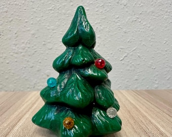 Könnte beinhalten: Eine kleine, kegelförmige Weihnachtsbaumfigur aus grüner Keramik mit glänzender Oberfläche. Der Baum hat geformte Zweige und ist mit vier kleinen, runden, durchscheinenden Kunststoffkugeln in den Farben Rot, Blau, Transparent und Bernstein verziert.