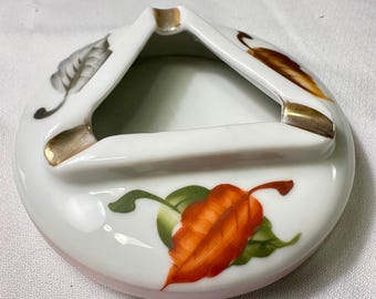 May include: White ceramic ashtray with a triangular center and gold-colored accents. The ashtray is decorated with hand-painted leaves in shades of orange, green, and gray. The ashtray has a circular base with a pink rim.