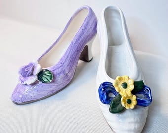 Könnte beinhalten: Zwei Schuhfiguren aus Keramik. Eine ist ein lavendelfarbener Pumps mit Absatz, einer violetten Rose und einem grünen Blatt. Die andere ist ein weißer flacher Schuh mit einer blauen Schleife, zwei gelben Blumen und grünen Blättern.