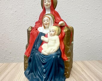 Könnte beinhalten: Eine Keramikfigur, die zwei auf einem goldfarbenen Thron sitzende Gestalten darstellt. Die größere Figur trägt ein rotes Gewand und ein weißes Kopftuch, während die kleinere Figur in ein dunkelblaues Gewand gehüllt ist und ein blondes Kind hält.