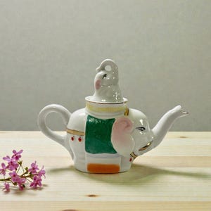 May include: White ceramic teapot in the shape of an elephant, with a small elephant lid. The teapot has a green blanket design, pink ears, and gold accents. The teapot is on a wooden surface.