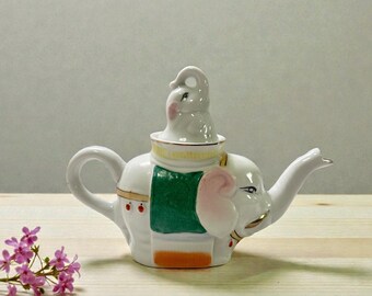 May include: White ceramic teapot in the shape of an elephant, with a small elephant lid. The teapot has a green blanket design, pink ears, and gold accents. The teapot is on a wooden surface.