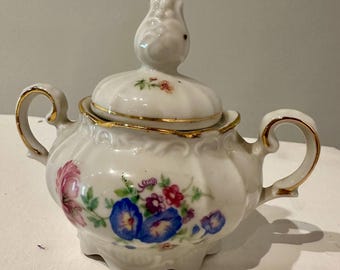 May include: A white porcelain sugar bowl with a lid, featuring a floral design in pink, blue, and red. The bowl has two handles and a gold trim. The lid is topped with a decorative finial.