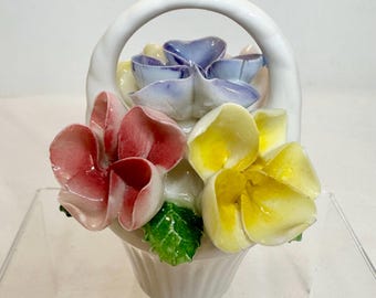 Könnte beinhalten: Eine weiße Keramik-Korbfigur mit einem hohen, gewölbten Henkel und einem Strauß aus modellierten Blumen. Das Arrangement besteht aus auffälligen rosa, gelben und hellblauen Blütenblättern mit grünen Blättern, alles mit einer glänzenden Glasur.