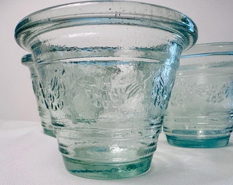 Könnte beinhalten: Ein Set aus drei hellblauen Glasübertöpfen mit breitem, ausgestelltem Rand und konischem Boden. Die Außenfläche weist ein strukturiertes, geprägtes Muster aus Blättern und Früchten auf, das den Mittelteil umgibt, gefertigt aus dickem, glattem Glas.