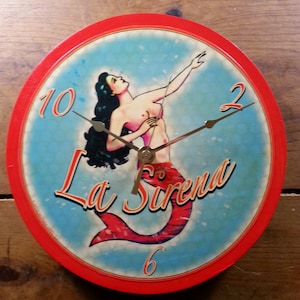 May include: Round wall clock with a red border and a blue face featuring a mermaid illustration. The clock face has the words "La Sirena" and the numbers 10, 2, and 6. The clock hands are black.