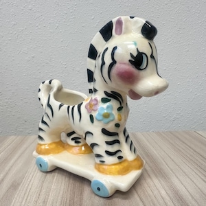May include: A ceramic zebra figurine planter featuring a glossy cream finish with black stripes, pink cheek accents, and small painted flowers. The zebra stands on a white platform base with four small light blue wheels and yellow hooves.