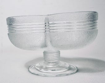 May include: A clear glass divided serving dish with a textured, pebbled surface. The bowl features two distinct compartments separated by a central divider, horizontal ribbed detailing along the sides, and a sturdy pedestal base with a circular foot.